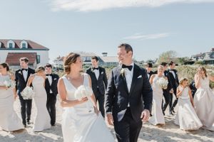 Wedding couple walking on sunny beach path surrounded by guests in formal attire.
