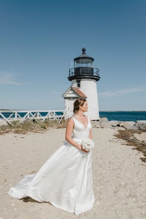 A white wedding dress stands out against a picturesque lighthouse backdrop on a sunny beach day.