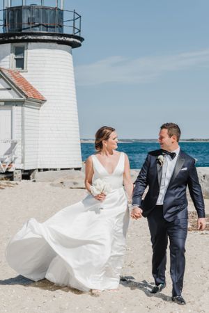 Couple walking hand in hand by white lighthouse on sandy beach with ocean backdrop.