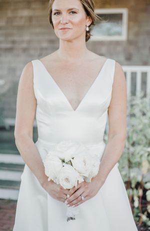 Classic white wedding gown with V-neck detail and white rose bouquet against rustic backdrop.