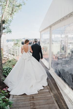 A wedding couple walks along a wooden deck beside a white tent pavilion with water views in the background.