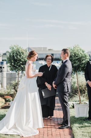 Outdoor waterfront wedding ceremony with potted trees and brick pathway.