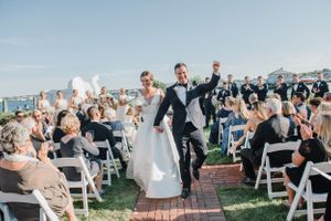 Newlyweds exit their outdoor wedding ceremony as guests cheer with white balloons.