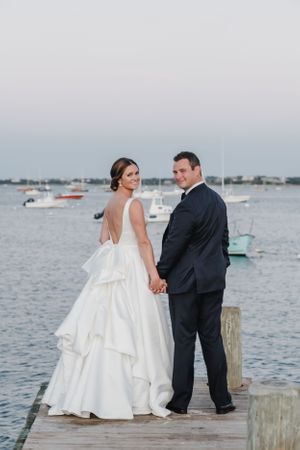 Wedding couple standing on wooden dock overlooking harbor with boats in background.