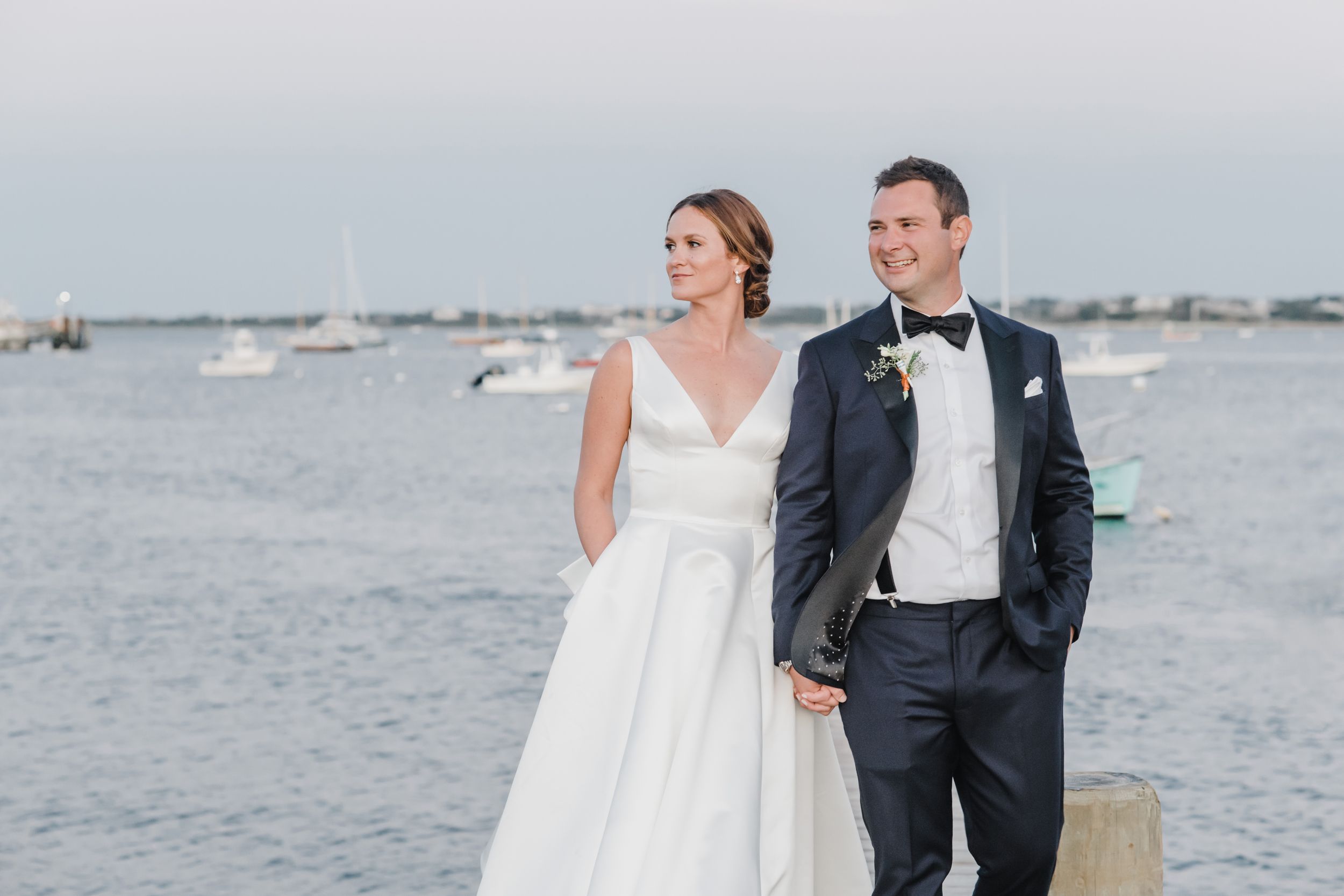 Wedding couple poses by the waterfront with boats in the harbor during golden hour.