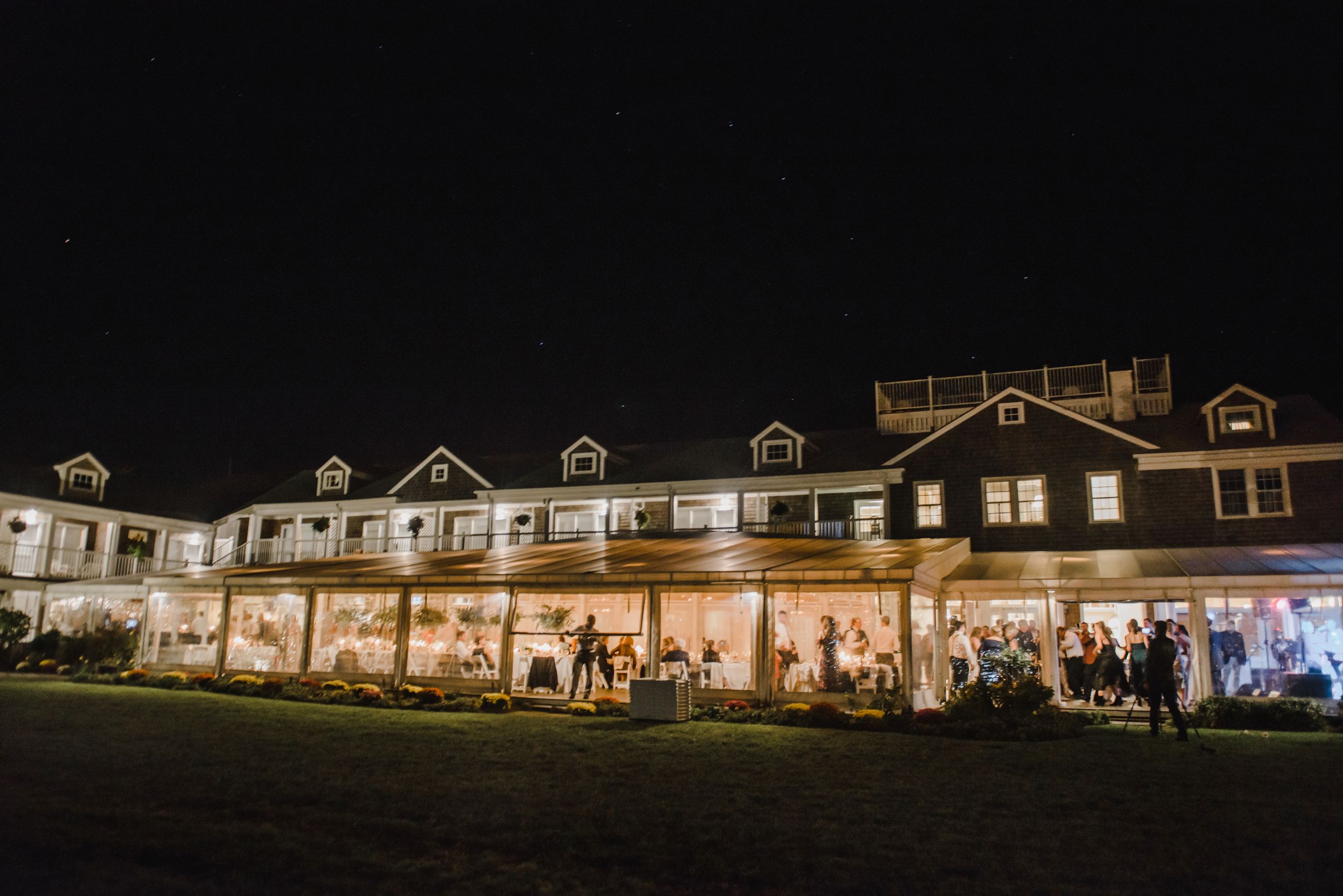 Elegant waterfront venue lit up at night with guests mingling on the porch during a wedding reception.
