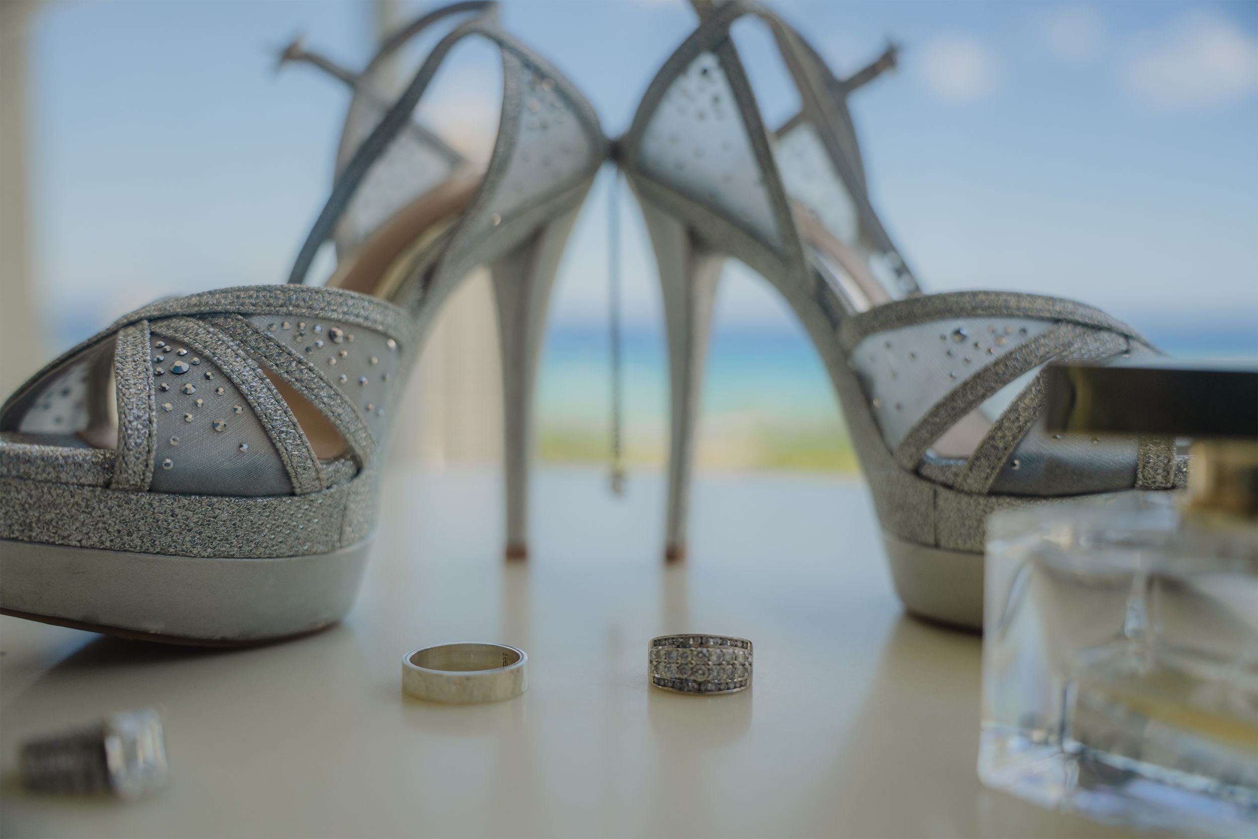 Silver glitter wedding shoes with crossed straps and wedding rings displayed on white surface with ocean view background.