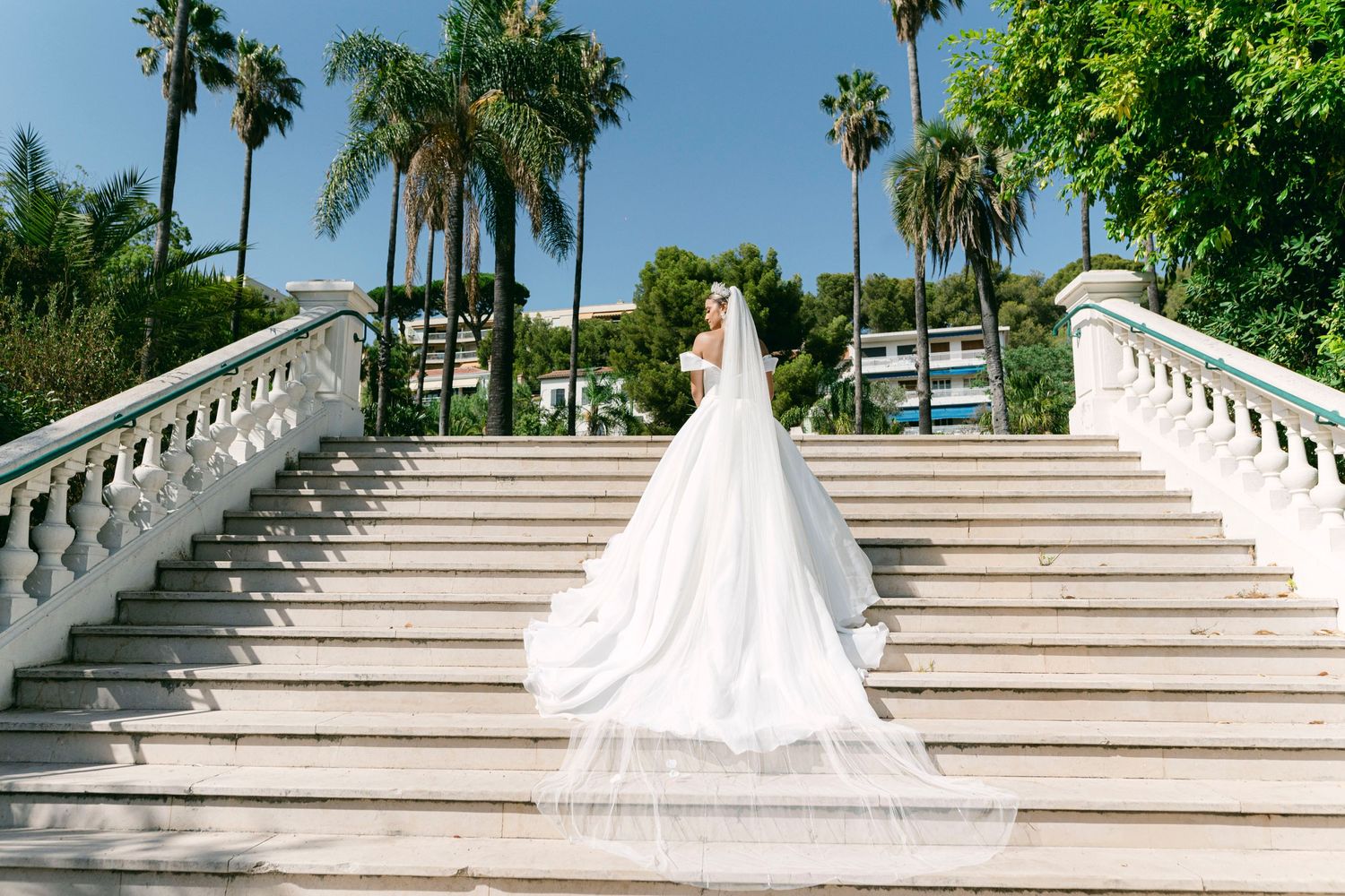 A bride in a flowing white gown with long train stands on grand marble stairs surrounded by palm trees at a tropical venue.
