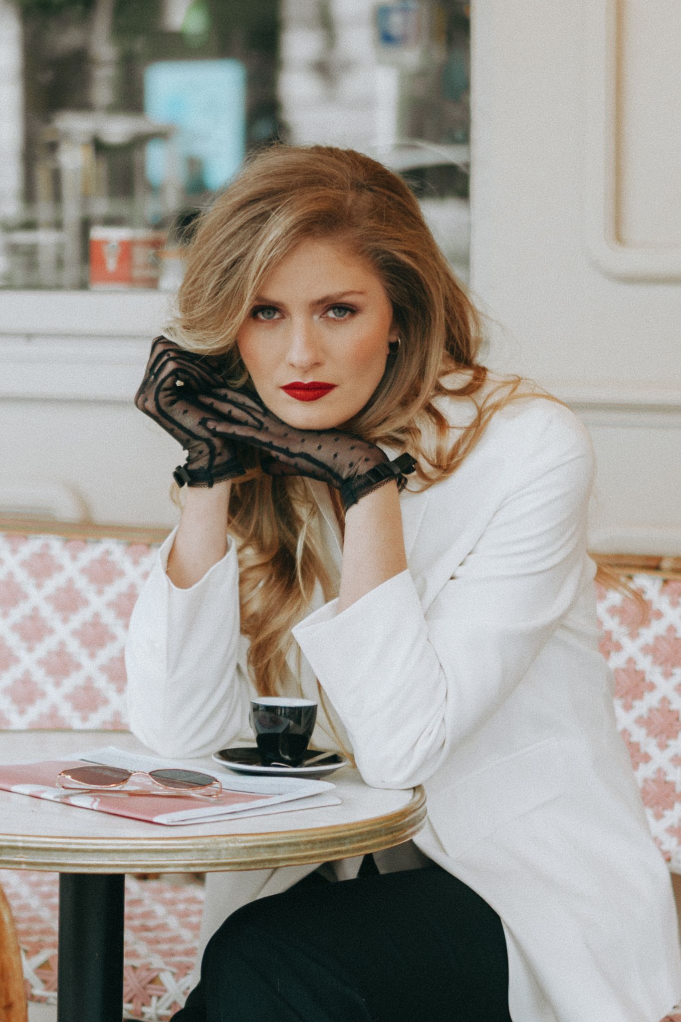 Fashionable woman in white blazer and black lace gloves sits at outdoor cafe table with coffee cup.