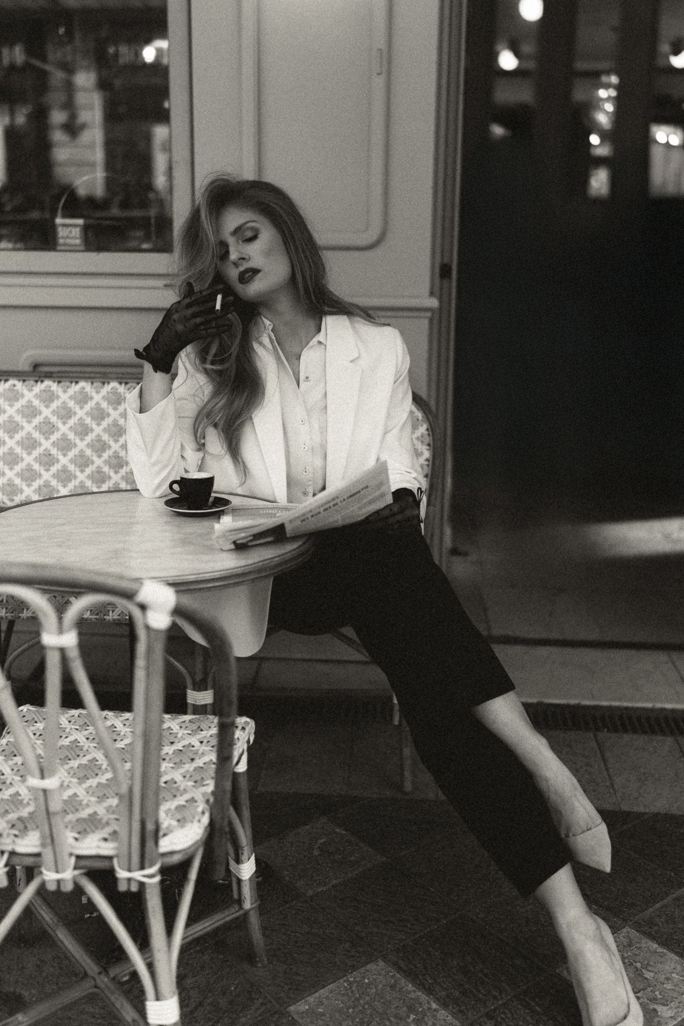 Élégante photo en noir et blanc d'un invité se relaxant à une table de café parisien portant un chemisier blanc et un pantalon foncé.