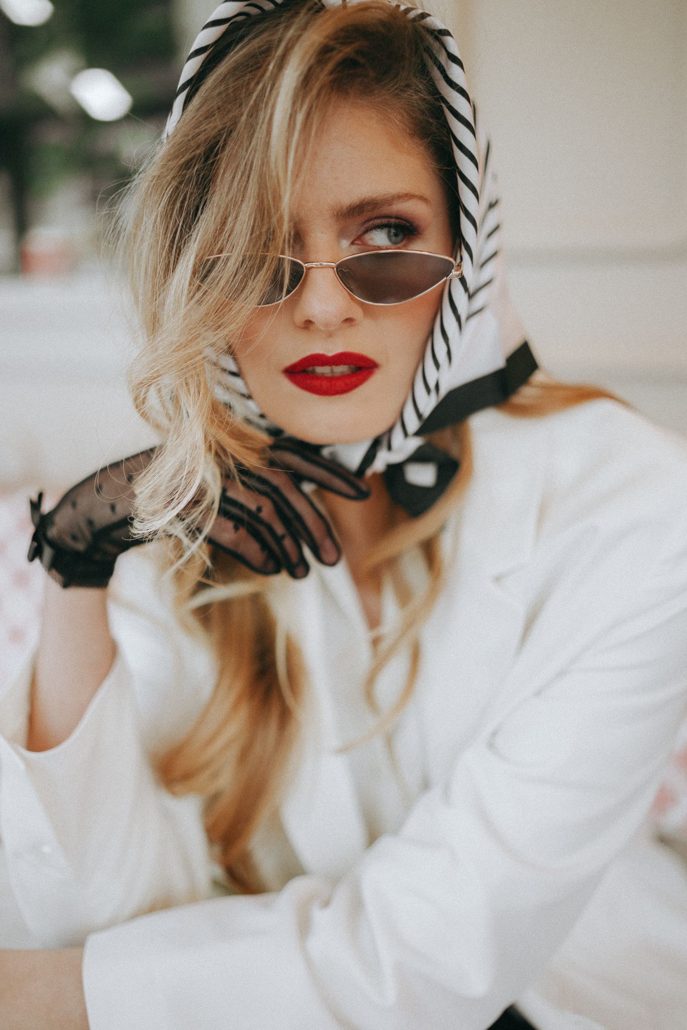 Portrait élégant avec foulard rayé, lunettes de soleil, rouge à lèvres rouge et pull blanc dans un éclairage naturel doux.