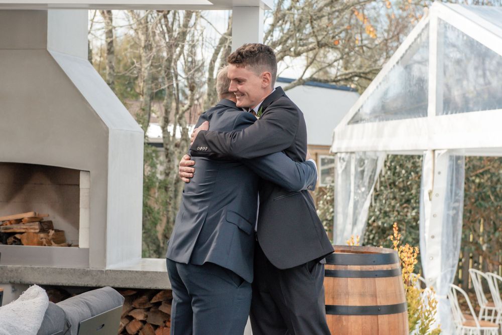 Two people in formal suits share an emotional embrace at an outdoor wedding venue with wooden barrels Cambridge New Zealand
