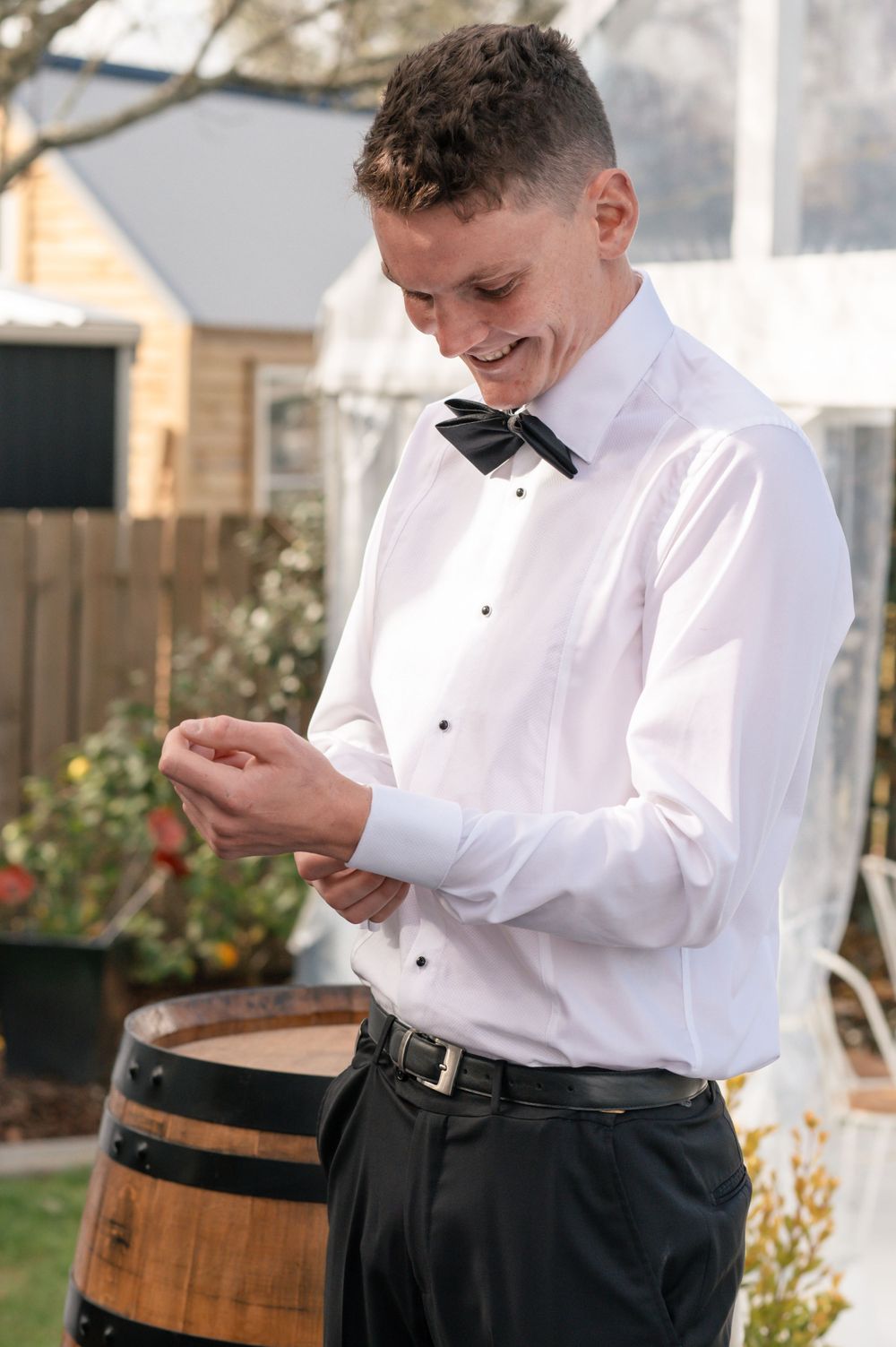 Groom in formal white shirt and bow tie smiles while looking down at his hand during outdoor wedding preparations Cambridge New Zealand