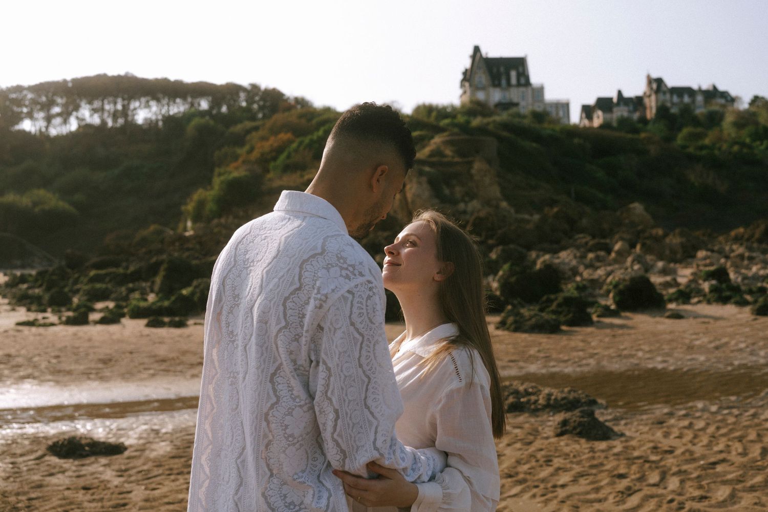 A romantic couple shares an intimate moment on a beach with a hilltop house in the background at sunset.