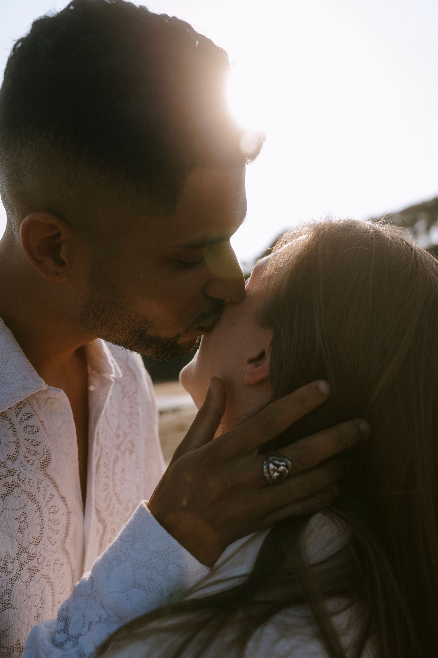 A close-up shot of a couple in white embracing tenderly against a sunlit coastal backdrop.