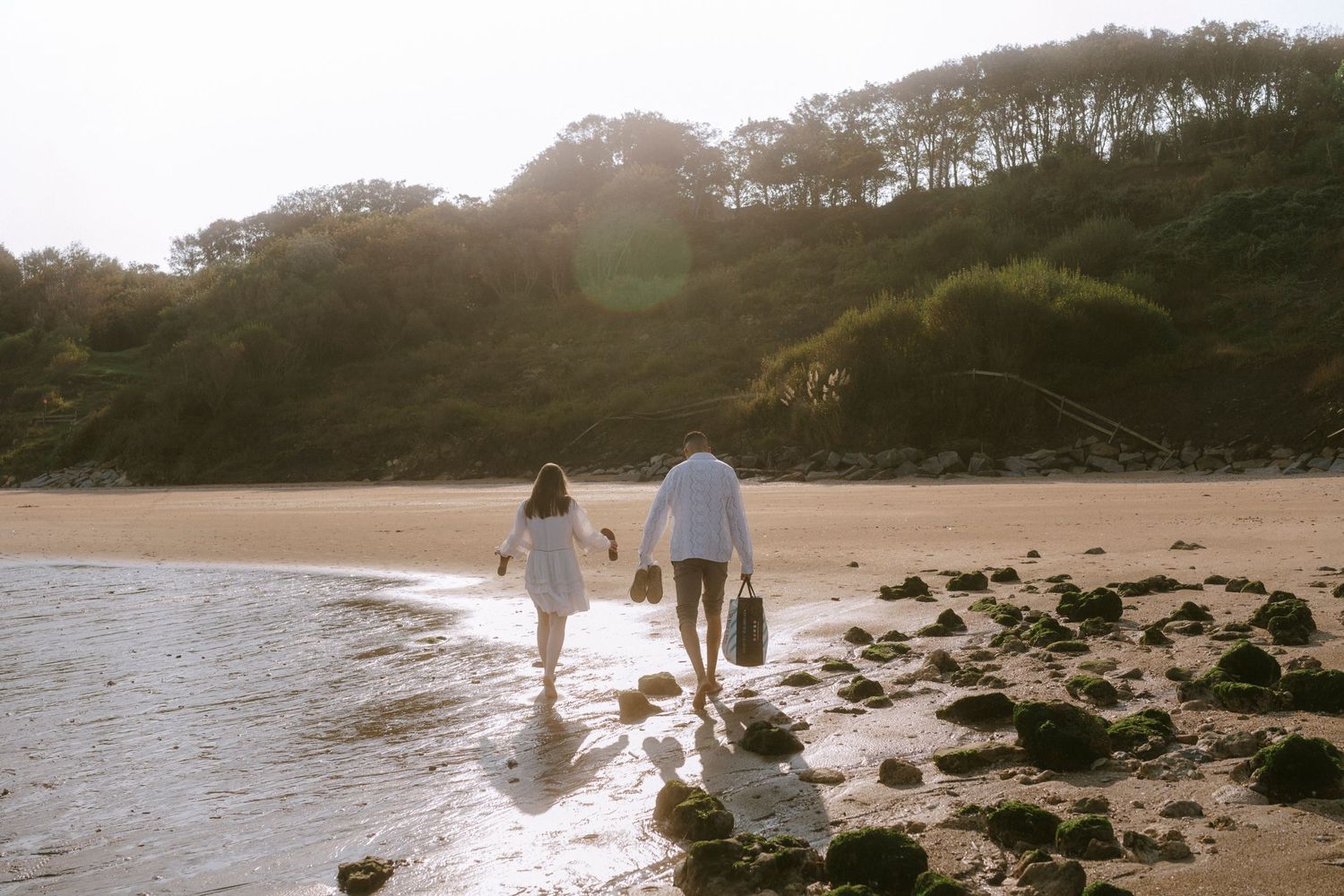 A couple walking hand in hand along a rocky beach shoreline at sunset.