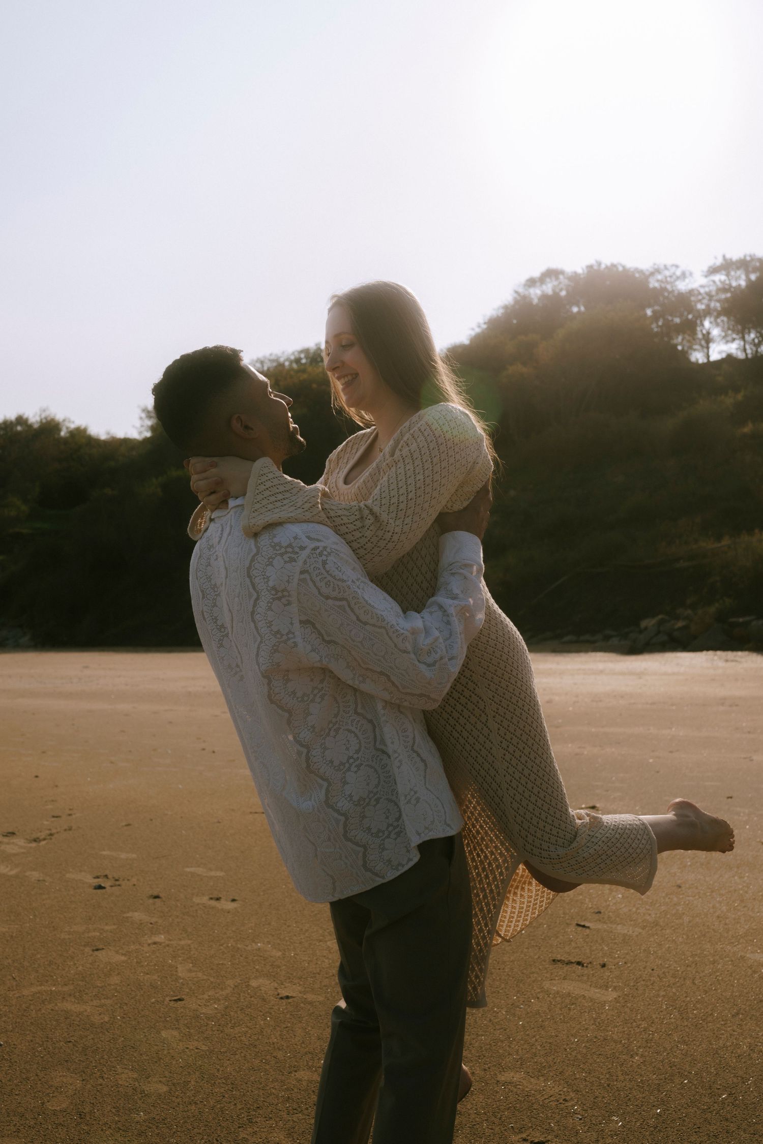 A couple in cozy sweaters embracing on a sandy beach during golden hour.