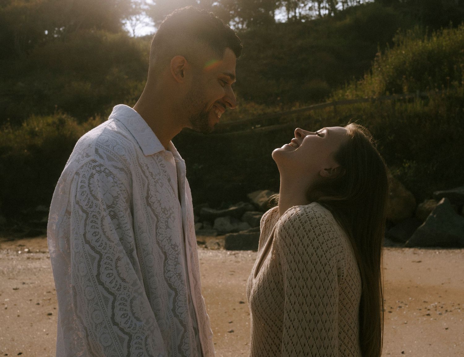 A couple in white knit sweaters sharing a joyful moment on a beach with hills in the background.