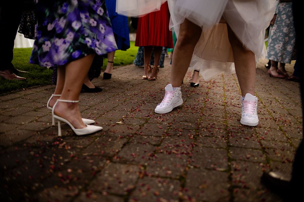 Close-up of wedding shoes and sneakers on brick pavement with scattered rose petals.