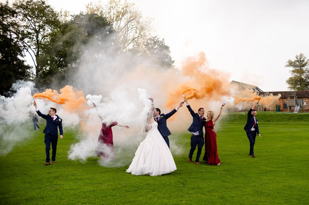 Wedding party creates colorful smoke bomb display on green lawn.