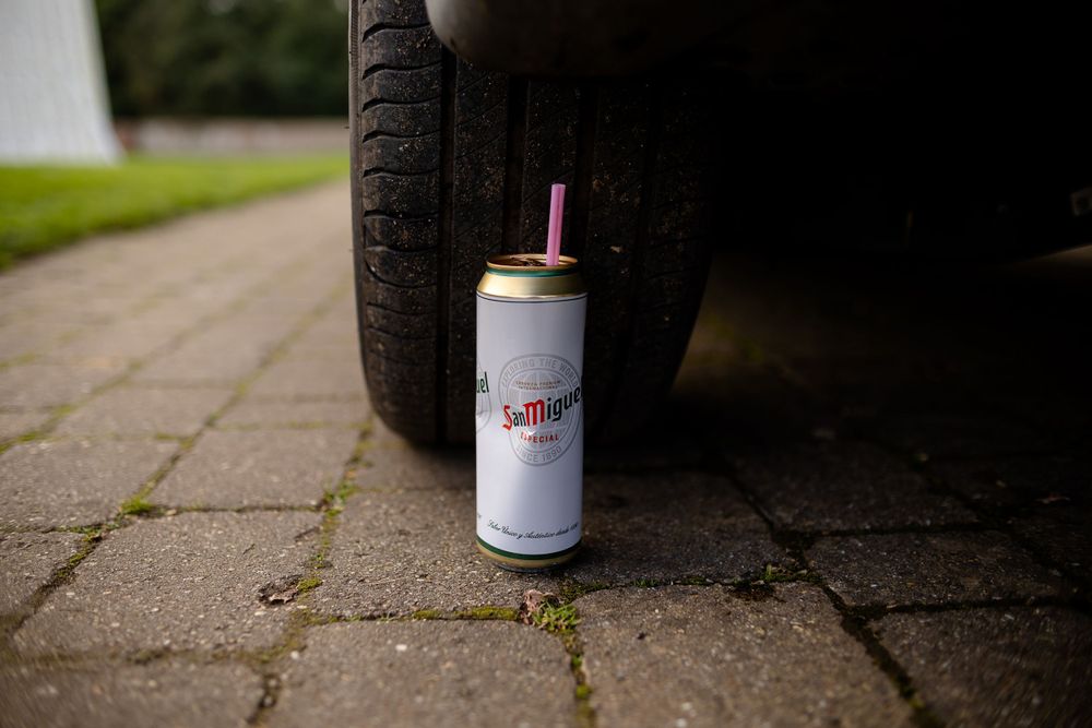A crushed beer can sits on pavement next to a car tire.