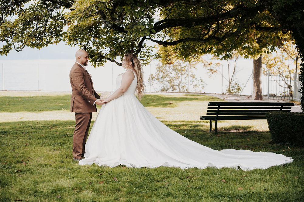 Bride and groom holding hands under a large tree in a park, wearing white gown and brown suit on their wedding day.