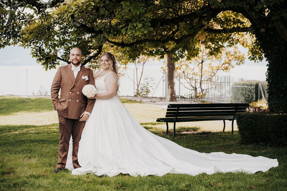 Bride in white ball gown and groom in brown suit pose under a tree on green lawn at outdoor wedding venue.