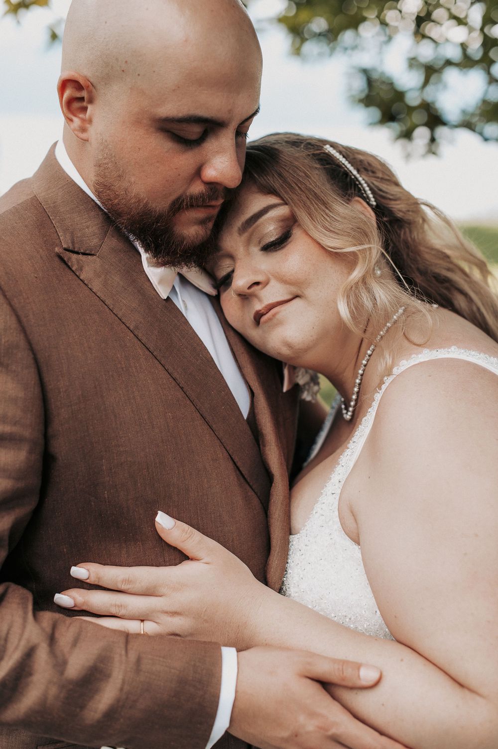 Bride and groom share a tender embrace, she rests her head on his chest wearing a white gown, he wears a brown suit.