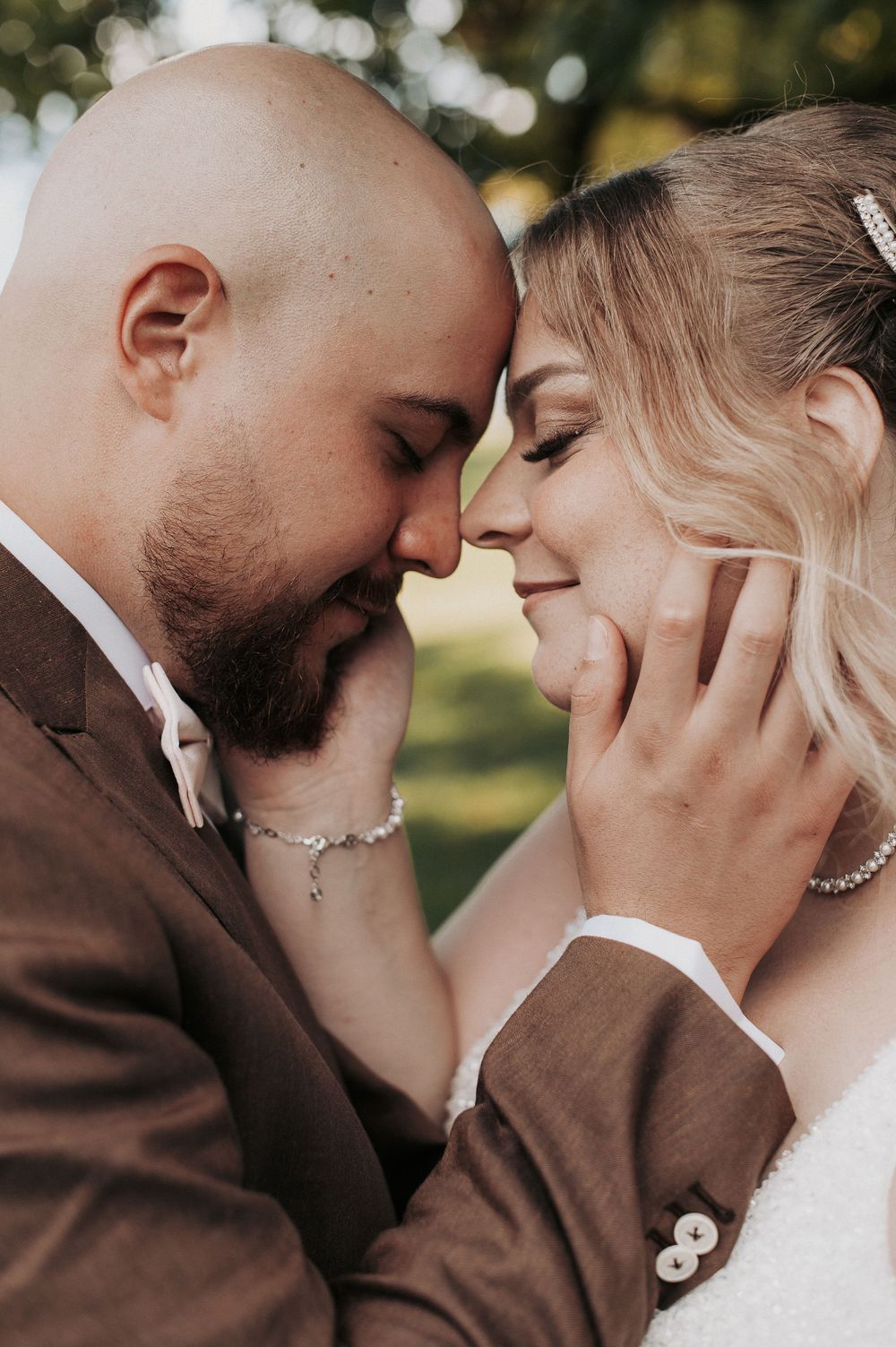 Bride and groom share an intimate forehead touch moment, groom in brown suit, bride in white gown with pearl accessories.