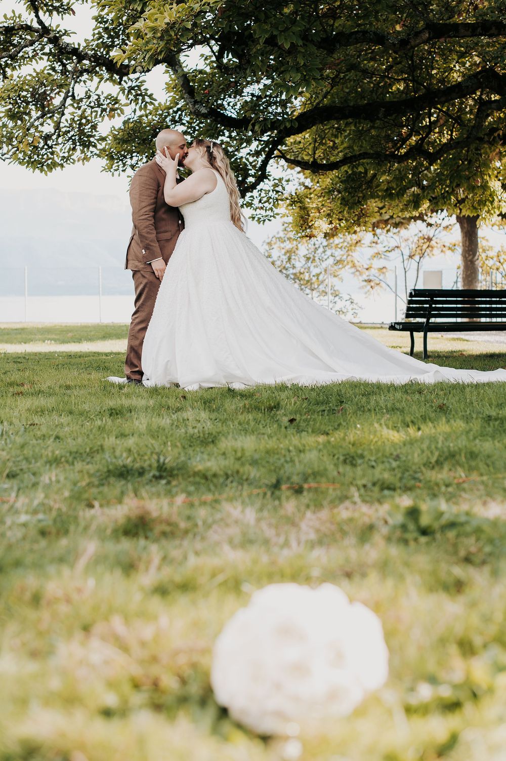 Bride and groom share a romantic kiss under a large tree in a park, brides white ballgown flowing in the breeze.