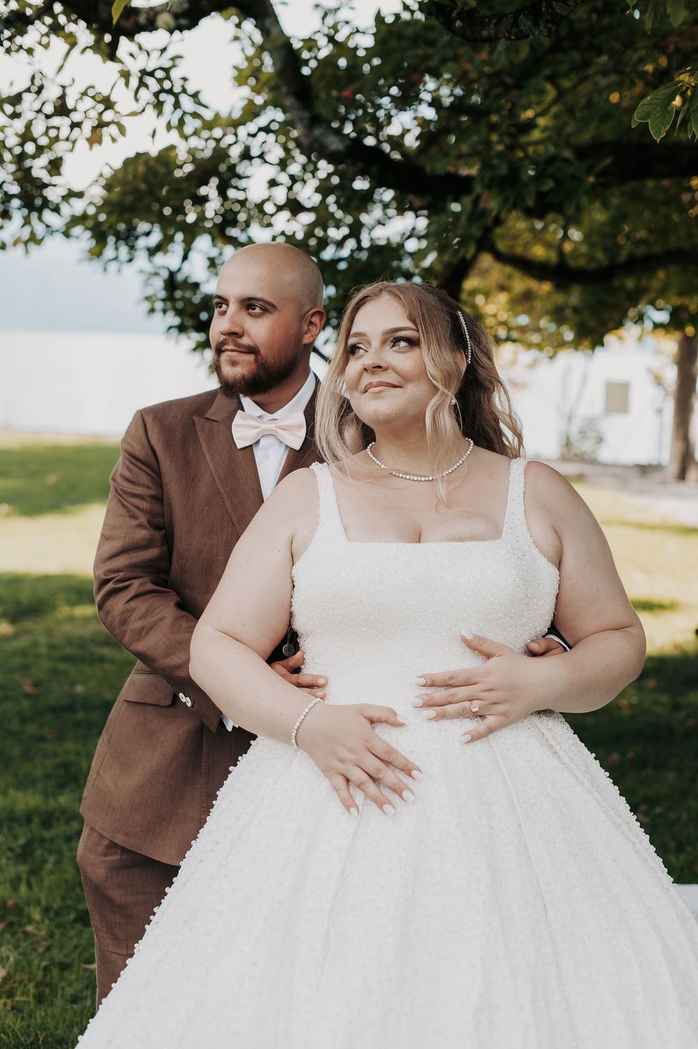 Bride in white gown and groom in brown suit embrace outdoors near a lake under a large tree.