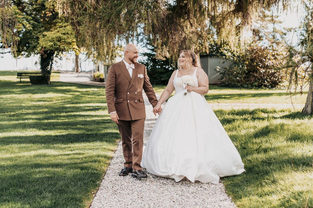Newlywed couple walks hand in hand along a garden path, groom in brown suit and bride in a white ballgown.