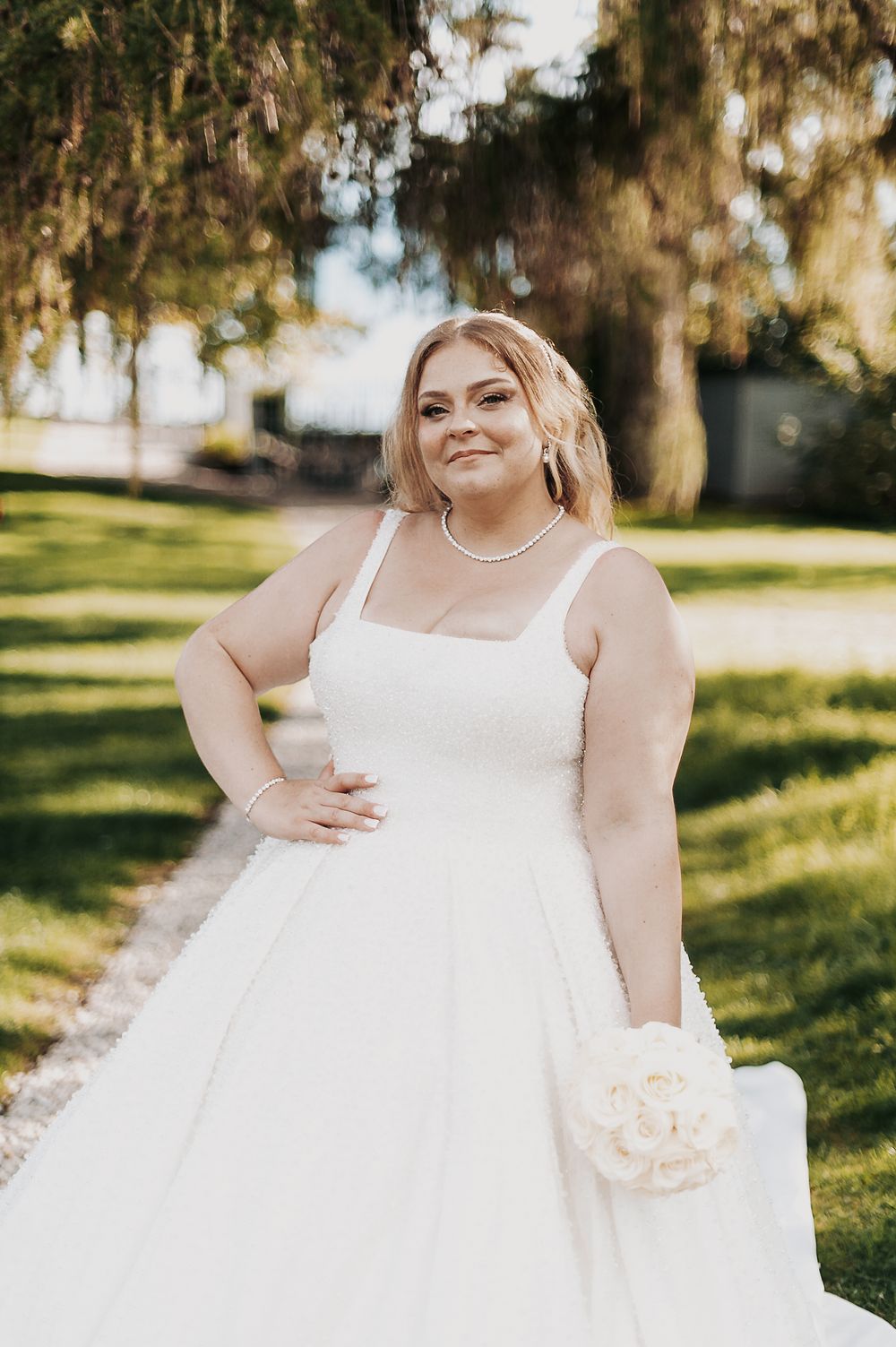 Smiling bride in white square-neck plus-size wedding gown holding bouquet outdoors in lush green garden setting.