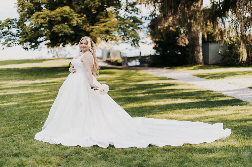 Bride in a stunning white ball gown with long train poses gracefully on lush green lawn outdoors.