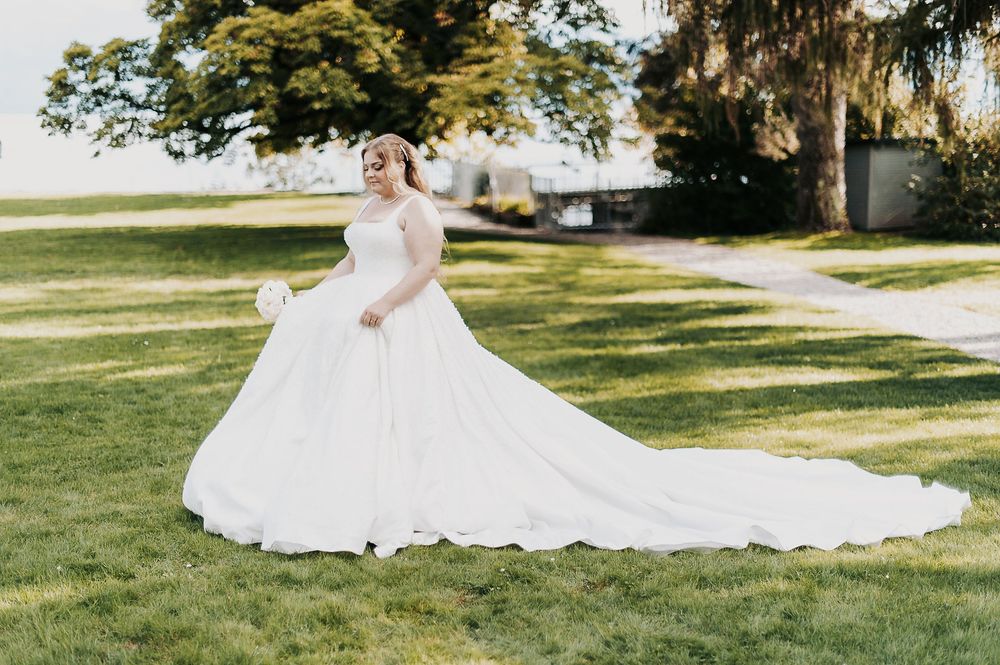 Elegant bride twirling in a white ball gown with flowing train on a green lawn surrounded by trees.