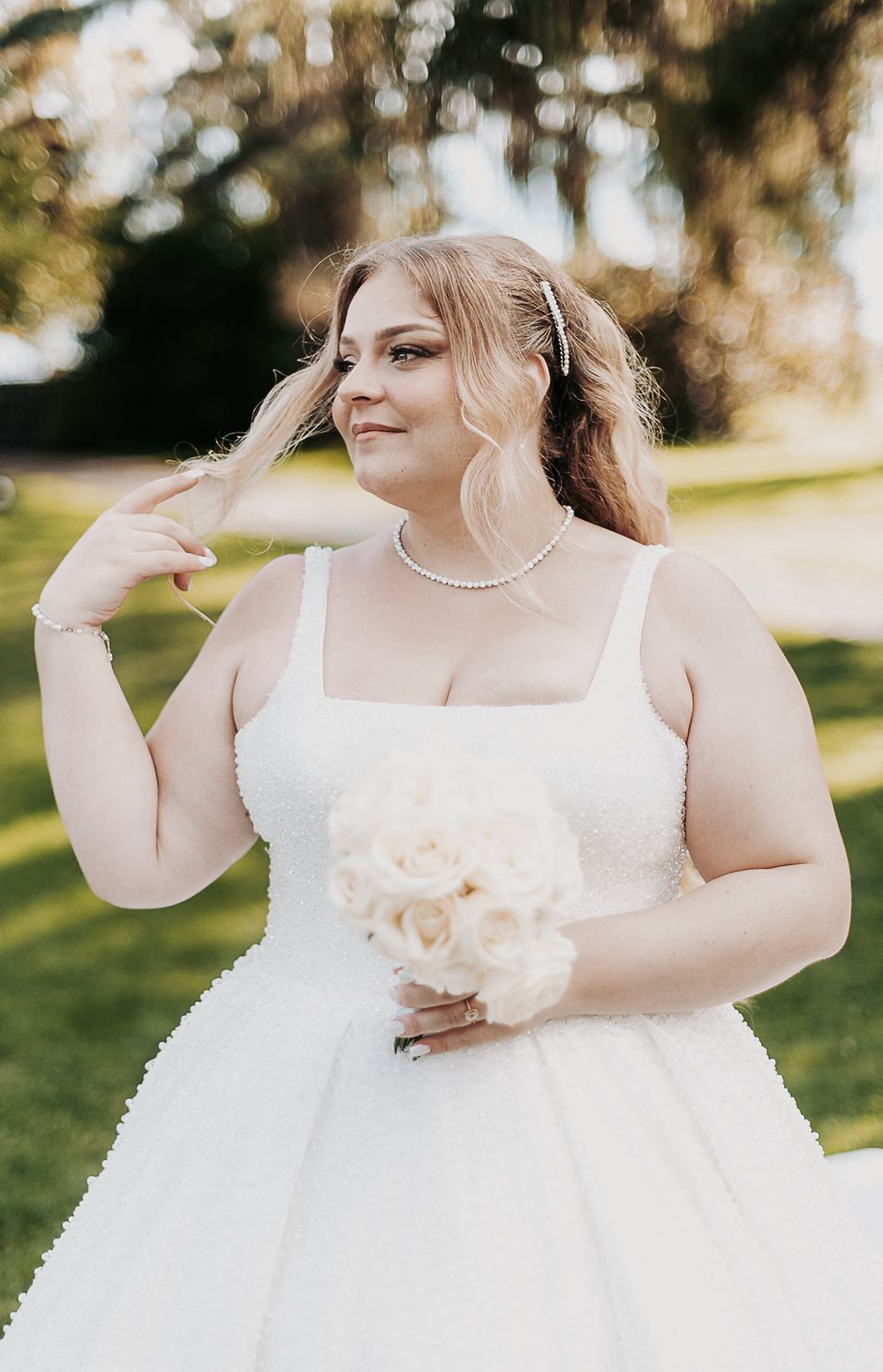 Plus-size bride holding white rose bouquet wearing pearl necklace and square-neck wedding gown outdoors.