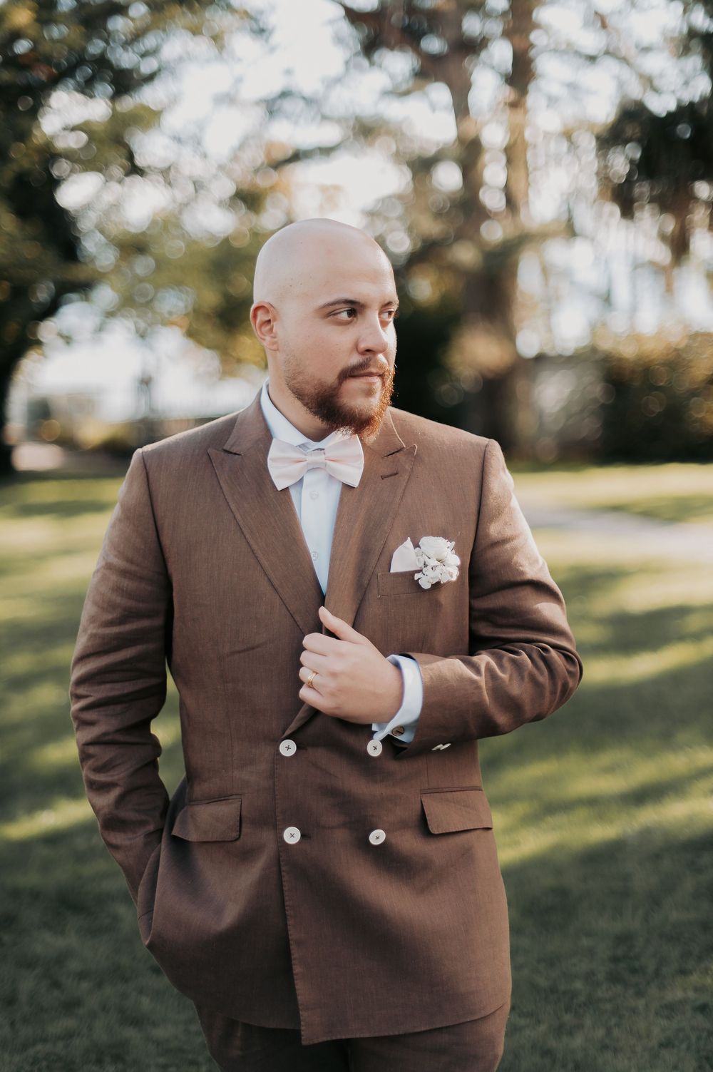 Bald groom with beard wearing brown double-breasted suit, white bow tie, and boutonniere, posing outdoors.