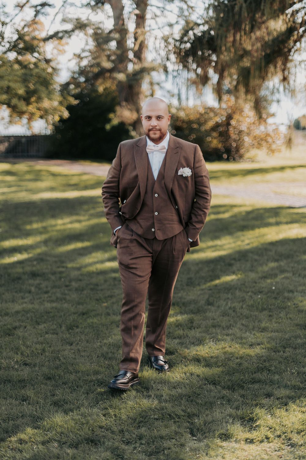 A groom in a brown three-piece suit with white pocket square stands on green grass outdoors.
