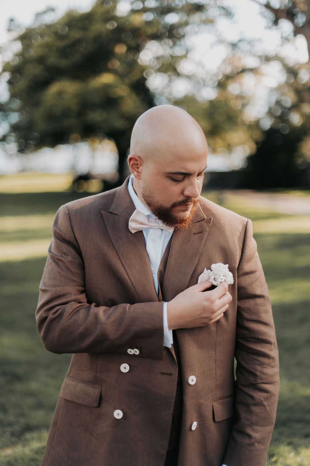 Bald groom with red beard adjusting white boutonniere on brown double-breasted suit with bow tie outdoors.