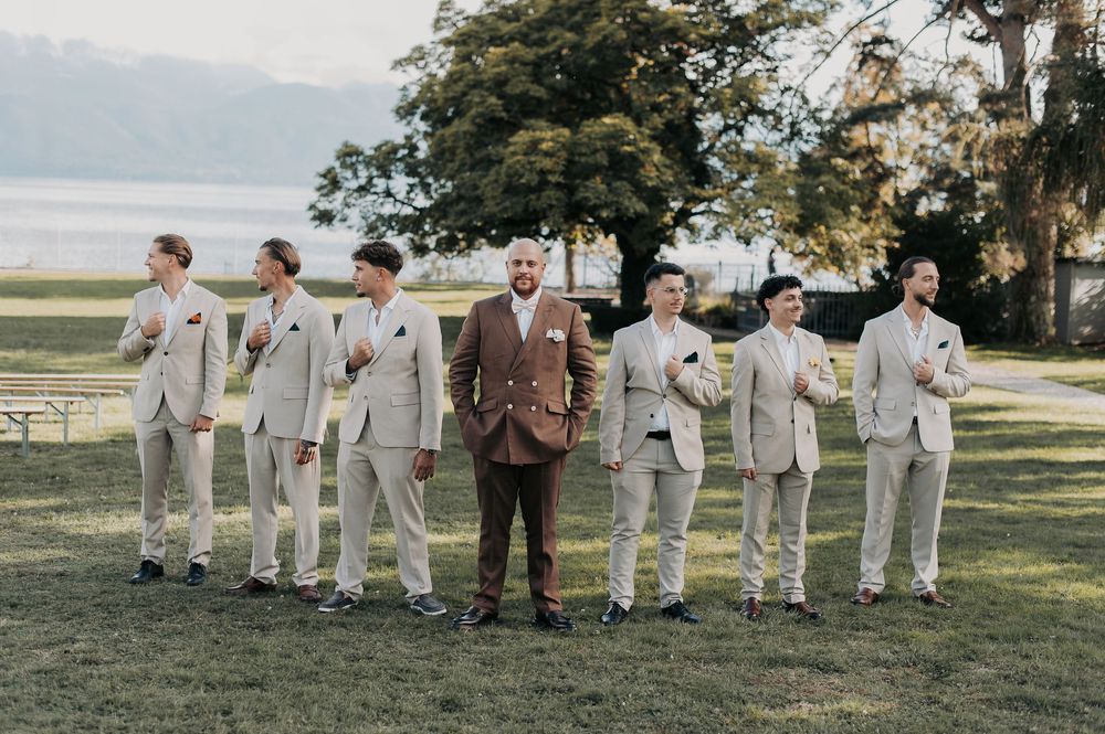 Groom in brown suit stands with six groomsmen in matching beige suits on green lawn with trees and water in background.