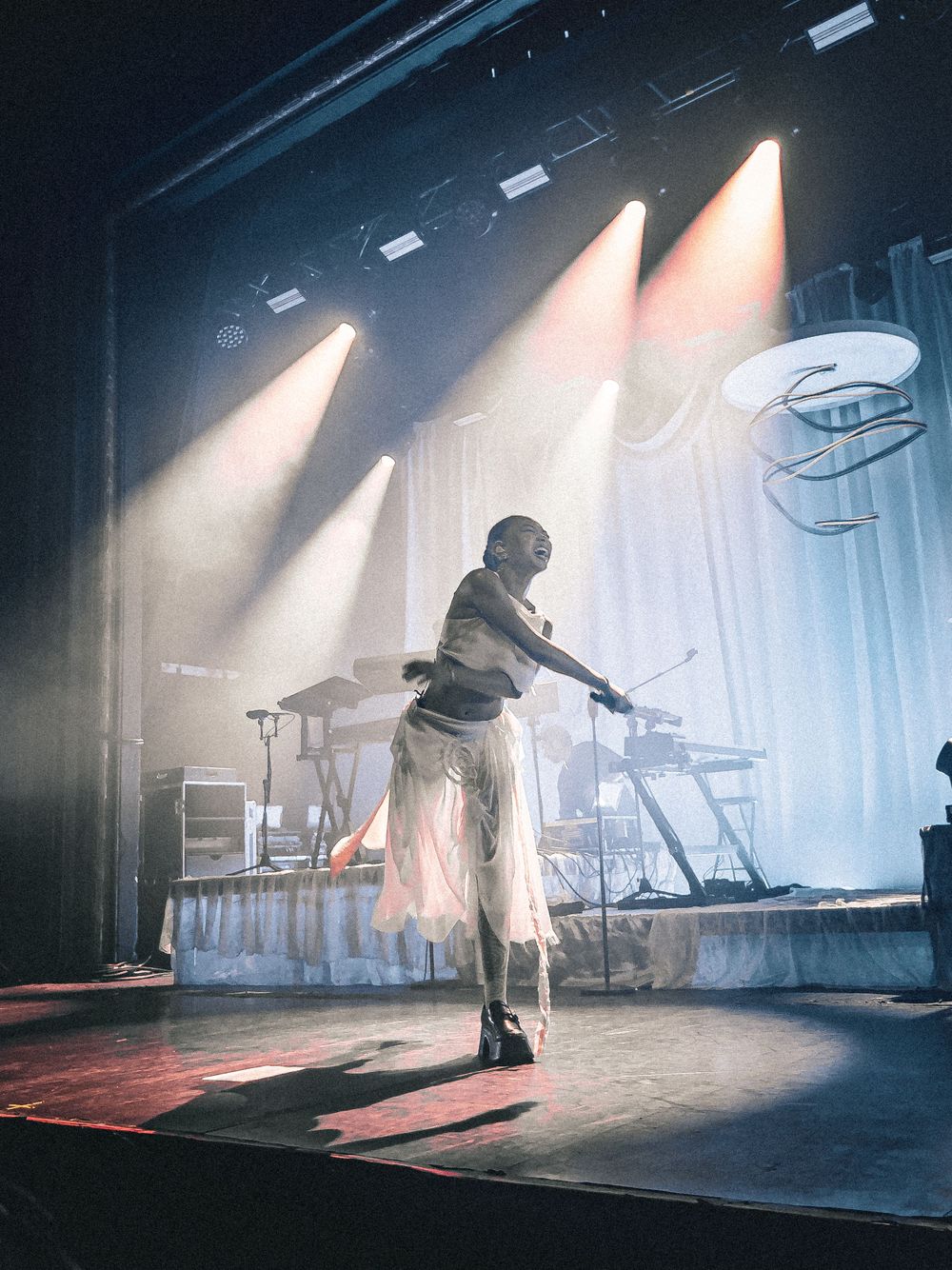 Performer in white outfit on stage with dramatic spotlights and electronic equipment.