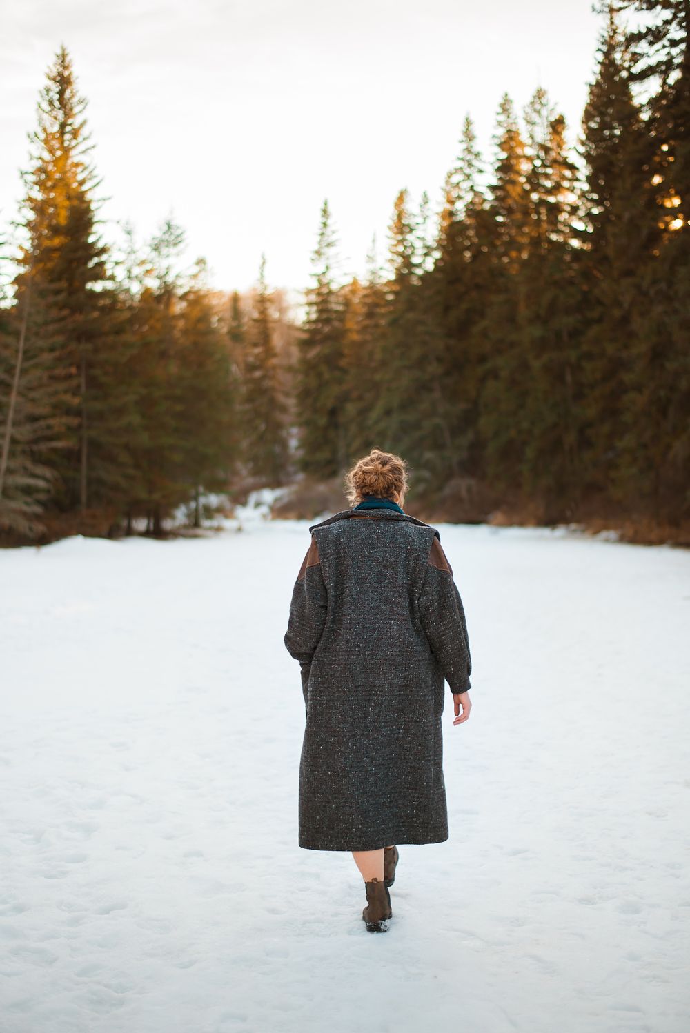 Person in long winter coat walking away on snowy path surrounded by pine trees.