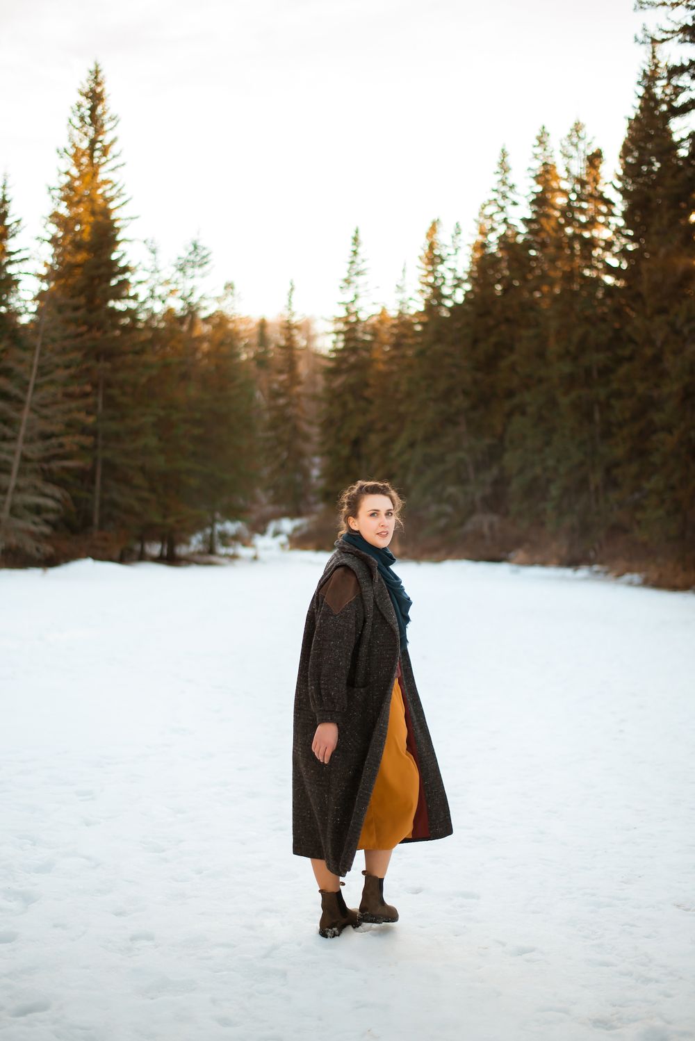 Winter fashion shot showing person in dark coat and mustard skirt against snowy forest setting.