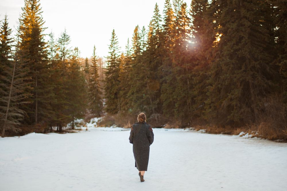 Person walking through a snowy forest path at sunset with evergreen trees in the background.