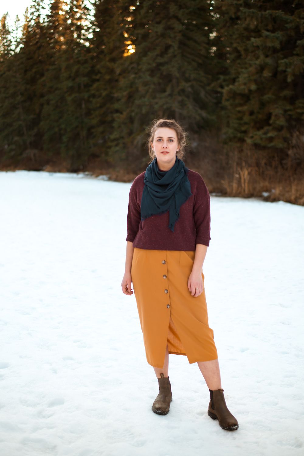 Person wearing burgundy sweater and mustard skirt stands in snowy forest setting.