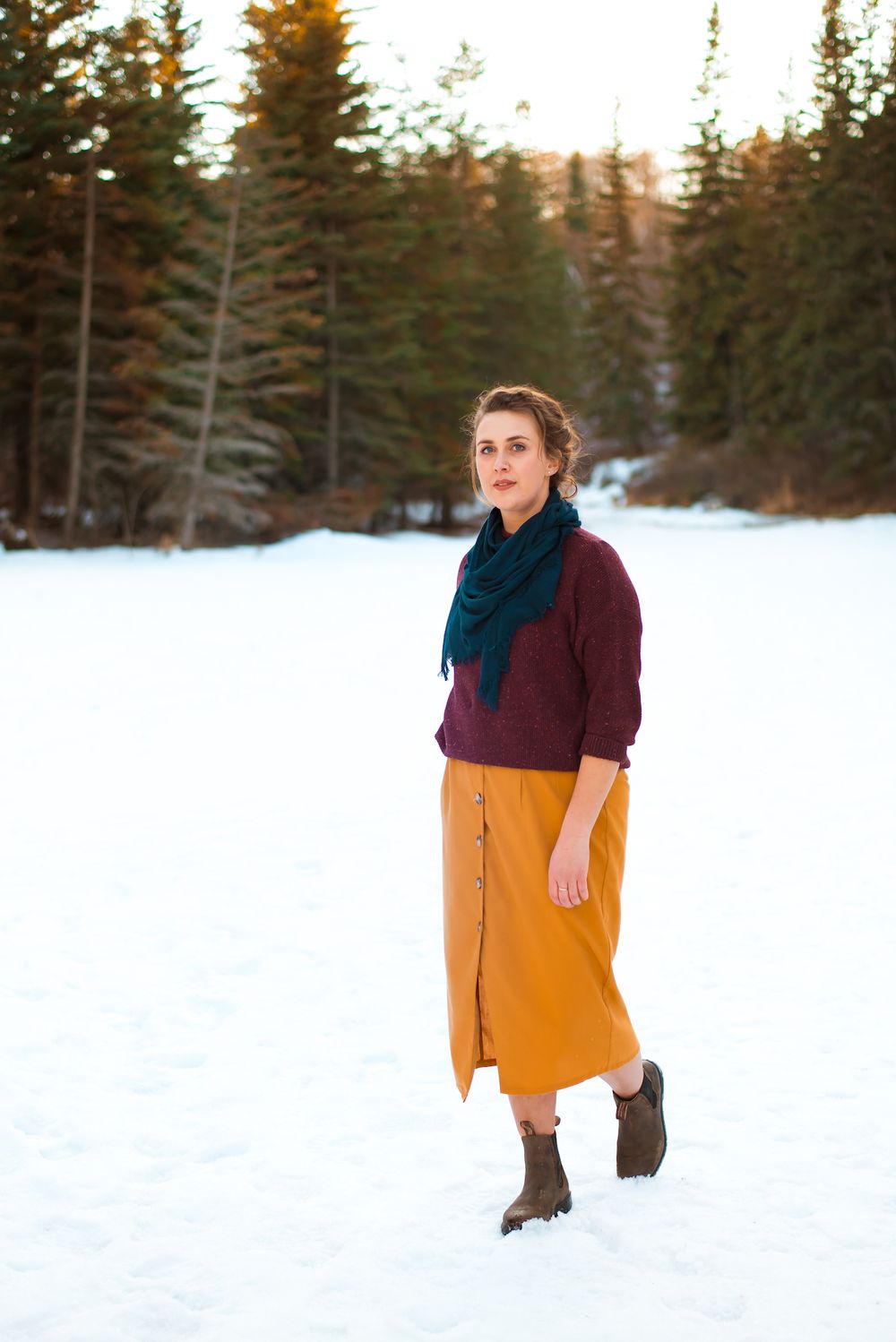 Winter fashion look with mustard skirt and burgundy sweater in snowy forest setting.