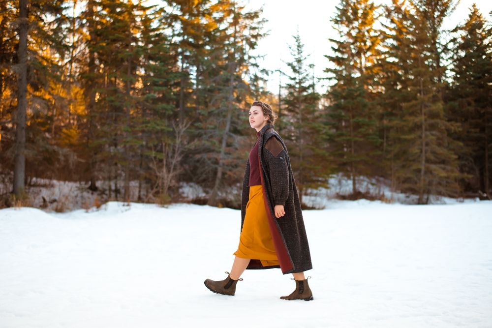 Figure walking in snow wearing long mustard skirt and dark coat among evergreen trees.
