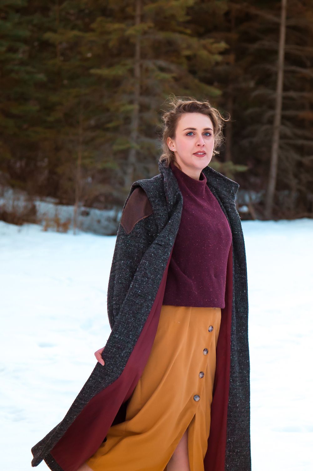 Winter fashion with burgundy sweater and charcoal coat against snowy forest backdrop.