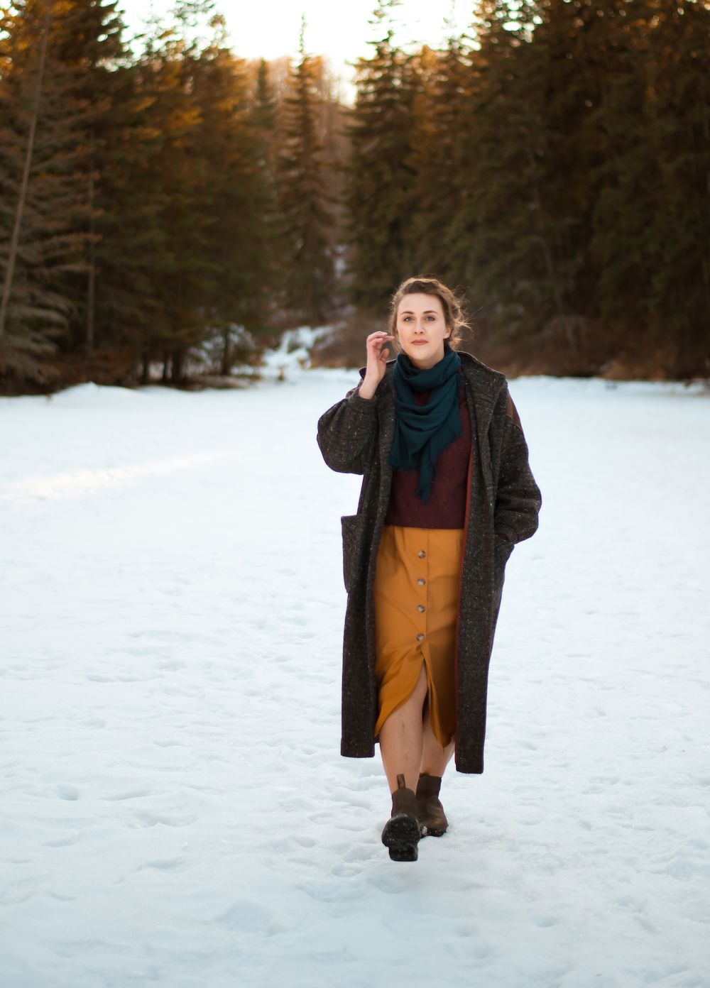 Person wearing long coat and navy scarf poses in snowy winter landscape with pine trees.