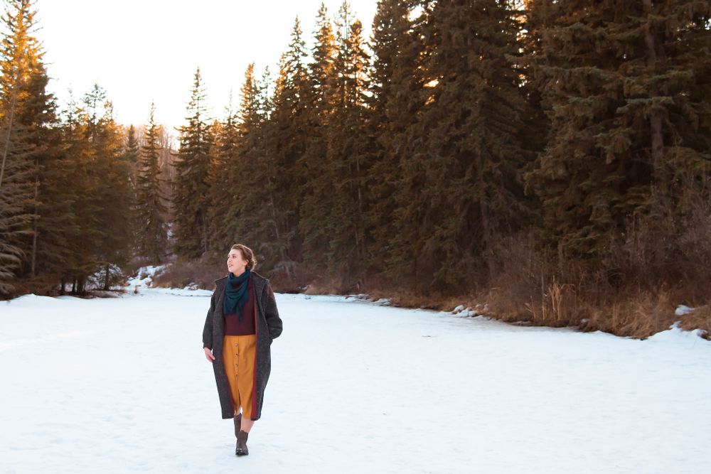 Figure in winter attire walking through snow-covered path with evergreen forest backdrop.
